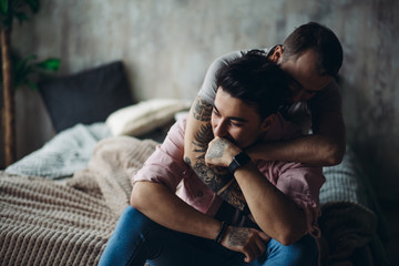 Happy gay couple embraced, joking and having fun in an intimate hug. Positive blue man embraces his friend, happy to be close to lover, sitting on bed at home, close up. Same-sex love relationship. - obrazy, fototapety, plakaty