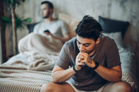 Young gay couple going through relationship problems. Pensive gloomy man dressed in t-shirt and underwear sits on bed with his hand leaning on chin with blurred image of his bridegroom on background