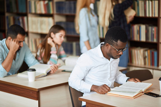 African american male college student in white shirt, wearing spectacles preparing for exams in the library, reading a book at the table.