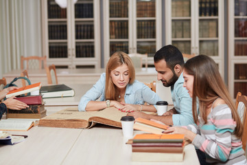 Mixed-race group of university students with a focus on blonde caucasian woman doing linguistic researches, turning over the pages of big old reference textbook at library.