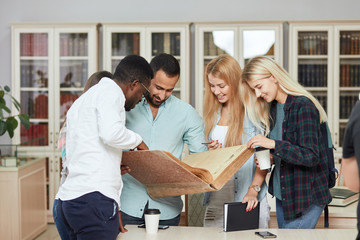 Cheerful Arabian unshaven male Tutor, dressed in casual blue shirt, holds in hands big old reference book, showing something to his diverse multiracial students, studying something.