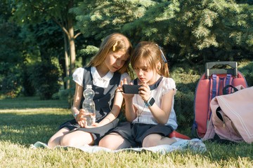 Two little schoolgirls using a smartphone. Children playing, reading, looking at the phone, in the park, golden hour. People, children, technology, friends and friendship concept