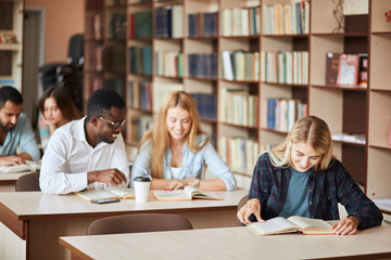 Young blonde woman writing notes with classmates studying in background. Students learning in college library.