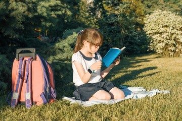 Little girl reading a book with a school backpack sitting on the grass in the park