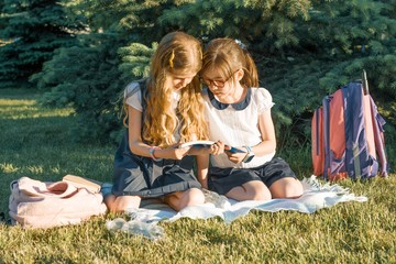 Two little girl friends schoolgirl learning sitting on a meadow in the park. Children with backpacks, books, notebooks