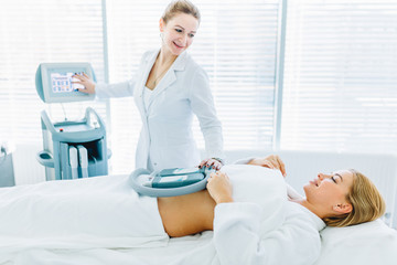 Young beautician in white uniform taking care of female client, lying on a table while getting a vibrant massage skin treatment combined with freezing effect in a healthy beauty spa salon.