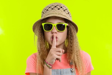 Portrait of teen girl in sunglasses and straw hat with a forefinger near the lips, showing silence sign, secret gesture, yellow studio background