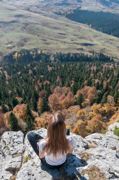 a young active girl sits on the edge of a rock, look at the Caucasus mountains.