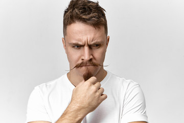Stress, problems and negative human emotions. Picture of handsome frustrated young male in white t-shirt posing indoors, frowning and touching his stylish beard, having worried uneasy look