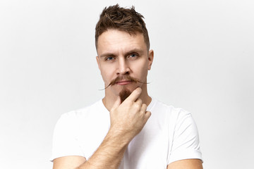 Fototapeta na wymiar Let me think. Pensive thoughtful young Caucasian male with stylish mustache touching his goatee beard while pondering, having deep in thoughts look. Body language and human facial expressions
