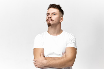 Attractive fashionable young bearded European hipster in white t-shirt crossing arms and looking...