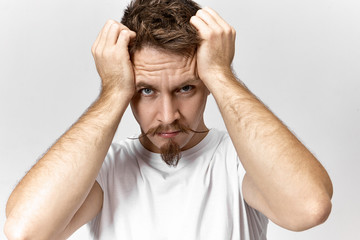 Close up shot of handsome young European man with stylish mustache and beard feeling stressed because of annoying noise. Bearded guy suffering form bad headache, squeezing temples. Pressure and stress