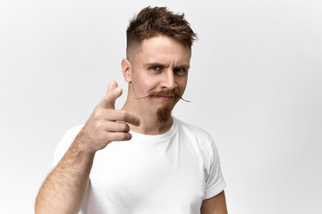 Signs, symbols, gestures and body language concept. Horizontal view of confident joyful young handsome guy with trendy hairstyle, mustache and beard smiling and pointing fore finger at camera