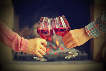 Man and woman hand toasting wine glasses in front of fireplace at home. Elderly couple have romantic dinner with red wine over fireplace background. Romantic winter night for couple in love. Toning.