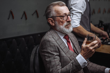 Bearded elderly businessman in stylish expensive suit drinking whiskey while getting a haircut by...