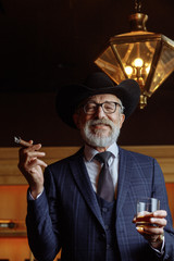 Stylish old-aged man in wide brimmed hat and rich dark blue mens suit smoking cigar indoor,...