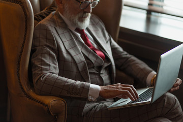 Fashionable focused elderly male writer wearing glasses and elegant clothes working on laptop,...