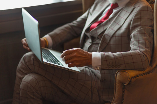 Senior bearded businessman in formal chic suit using laptop for work while sitting in big leather arm-chair in his office.