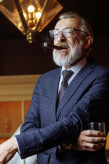 Successful old-aged european businessman spending night in luxury restaurant, smoking cigar and drinking alcohol beverages, looking at camera over bar counter background.