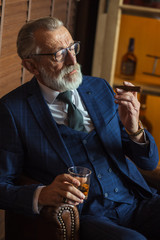 Senior cute business man with cigar and whisky. Gray hair and beard wearing blue tailored suit and tie, relaxing at fashionable restaurant lounge