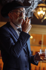 Big boss in retro style. Serious senior man in wide-brimmed hat and tailored suit, smoking cigar and looking at you while standing against pub interior.