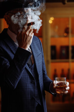 Concept Of Old Age, Wealth, Prosperity And Experience. Prosperious Confident Aged Man With Grey Beard And Spectacles, Wearing Big Dandy Hat Smoking Cigar And Sipping Cognac In Luxury Interior