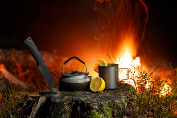 A mug, a teapot, a cut lemon and a knife stand on a stump in the forest at night. A bonfire with...