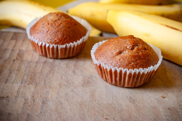 Closeup photo of the banana bread muffin in white paper cup and the bananas on wooden table background. The homemade easy bake oven cake.
