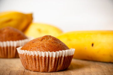 Closeup photo of the banana bread muffin in white paper cup and the bananas on wooden background. The homemade easy bake oven cake.