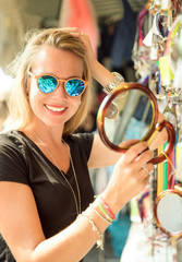 junge hübsche blonde frau auf dem markt in der sommer sonne in italien bei ihrer auslands reise. sie schaut sich sonnenbrillen, t-shirts, souveniers und taschen am markstand an und 
