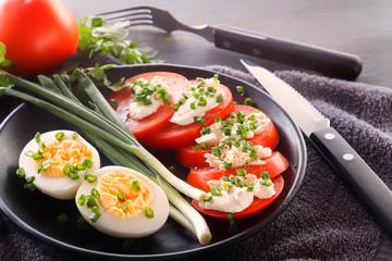 Half boiled eggs with tomatoes, green onions, sprinkled with greens in a black plate on a gray wooden table. Close-up