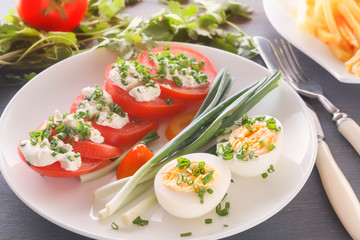 Half boiled eggs with tomatoes, green onions sprinkled with greens in a white plate on a gray wooden table. Close-up