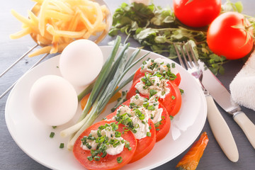 Sliced tomatoes with cheese and herbs on a white plate with onions, sweet peppers, eggs and french fries. Close-up