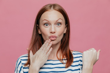 Close up shot of attractive woman raises eyebrows, pouts lips, keeps hand near mouth, has brown hair, surprised by something, models over pink background. People, facial expressions and beauty concept
