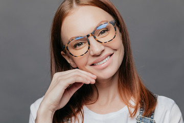 Close up portrait of pretty female model with European appearance, smiles gently, glad to recieve good comments from boss, wears spectacles, keeps hand near face, isolated over grey wall, tilts head