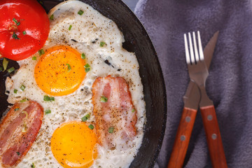 Fried eggs with bacon and tomatoes on an old cast iron pan and cutlery on a gray table. Close-up. Top view
