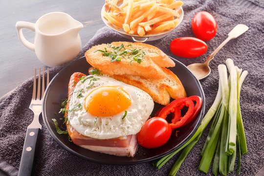 Fried egg with bacon in a black plate with fried pieces of bread, greens tomatoes, a jug of milk and French fries on a gray wooden table. Close-up