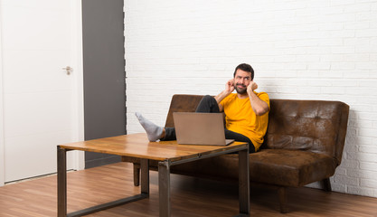 Man with his laptop in a room covering ears with hands. Frustrated expression