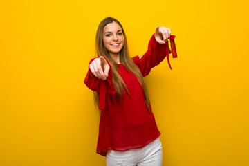 Young girl with red dress over yellow wall points finger at you while smiling