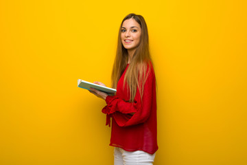 Young girl with red dress over yellow wall holding a book and enjoying reading
