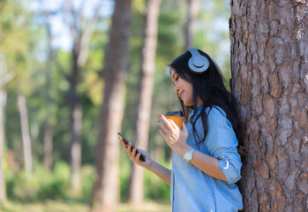 Young woman listening music in modern headphones communicating online  with cup of coffee on hand in the pine forest.