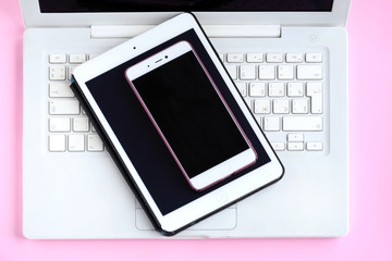 Tablet and mobile phone with black screens on the  white laptop keyboard. Electronic touch device with selective focus on pastel pink background. Mobility wireless web office with digital equipment.