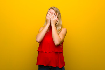 Young girl with red dress over yellow wall covering eyes by hands and looking through the fingers
