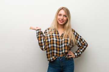 Blonde young girl over white wall holding copyspace imaginary on the palm to insert an ad