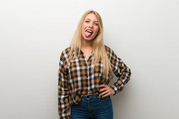 Blonde young girl over white wall showing tongue at the camera having funny look