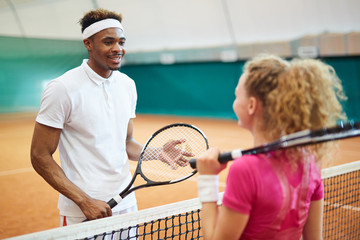 Two young intercultural tennis players with rackets standing by net before game