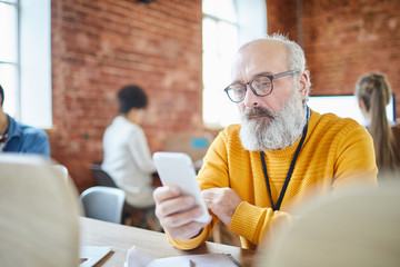 Bearded mature businessman reading notification or scrolling in smartphone by workplace