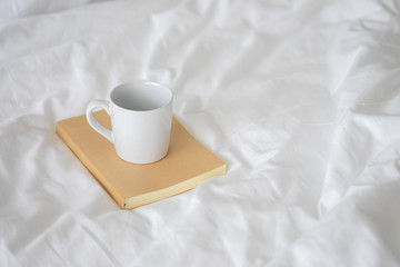 Fototapeta na wymiar White ceramic coffee mug placed on Notebook brown cover. Put on bed a blanket and white linen. Keep your knowledge clean and comfortable. Perfect for relaxation.