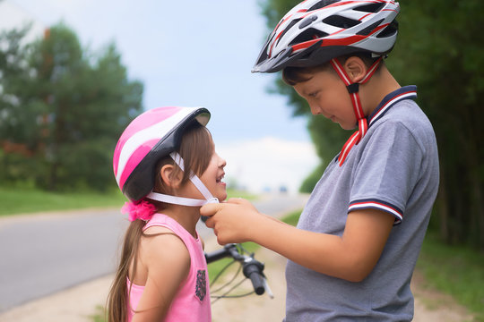 Children learn to ride bicycle in a park on summer day. Teenager boy helping preschooler girl to put on safety helmet. Active outdoor sport for child.