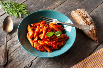 Green plate with bolognese macaroni, mint leaves, bread and spoon and fork on white rustic table top view
